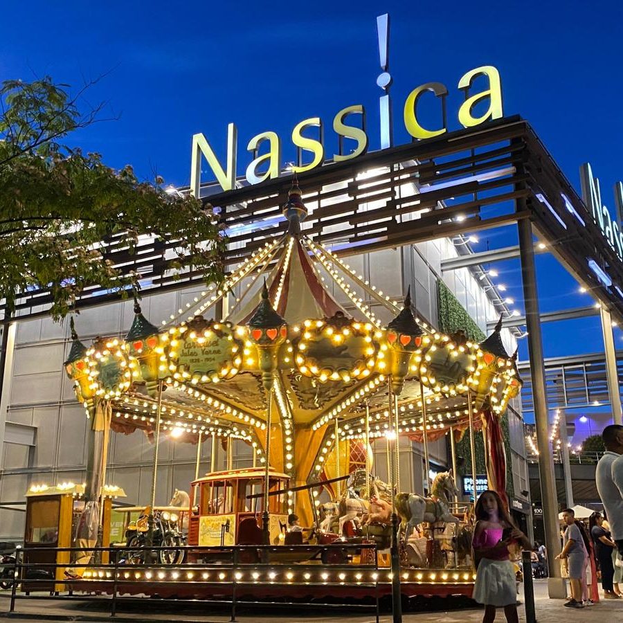 El carrusel Julio Verne Carrusel iluminado en un centro comercial con el letrero "Nassica" al fondo.